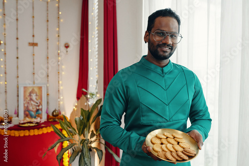 Man in green kurta smiling while holding plate of traditional festival sweets. Offering festive food in front of home altar during celebration near window photo