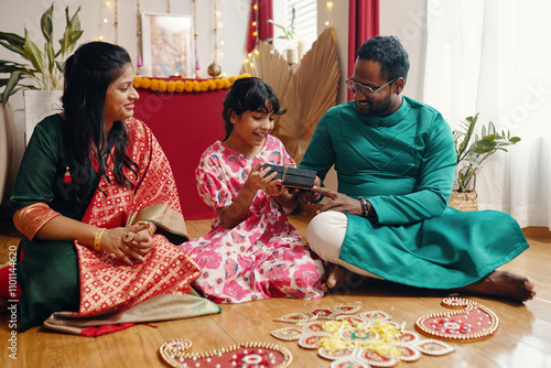 Family dressed in traditional attire, sharing a joyful moment while sitting together in a decorated room, holding and presenting gift boxes. Happy child looking excited photo