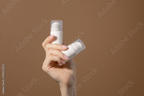 Woman's hand holds white unmarked bottles for cosmetics. Mock up of container for serum, lotion, cream, retinol. Light brown background with copy space