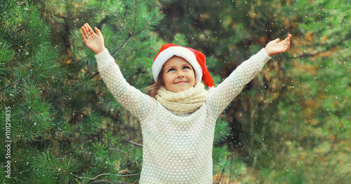 Happy smiling little girl child in santa red hat, snow in forest on Christmas tree background