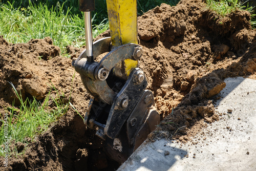 Mini excavator digs a narrow trench in soil for laying communications close-up. Mini bucket of crawler machine. Earthworks with heavy special equipment. Digging machinery works at construction site