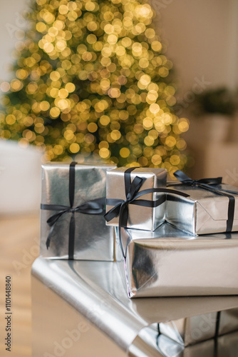 Close-up of a stack of Christmas gifts wrapped in silver paper in front of an illuminated Christmas tree