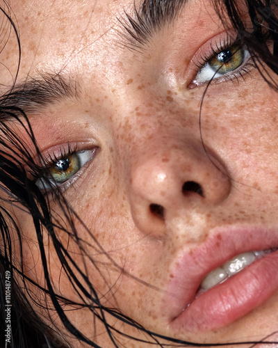 Intimate close-up of a freckled woman with glowing skin, minimal makeup, and wet hair. The artistic composition highlights her natural beauty and raw elegance