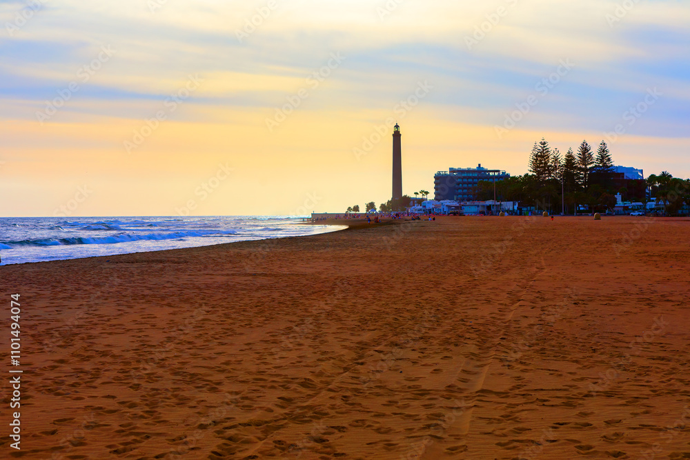Maspalomas lighthouse and beach at twilight, Gran Canaria, Canary Islands. Sandy beach during sunset with a tall lighthouse. Beach is with footprints in the sand, and the ocean waves against shore