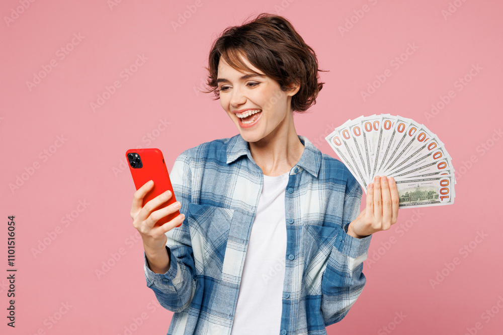 Young woman wear blue shirt white t-shirt casual clothes hold in hand fan of cash money in dollar banknotes use mobile cell phone isolated on plain pastel light background studio. Lifestyle concept. Young woman wear blue shirt white t-shirt casual clothes hold in hand fan of cash money in dollar banknotes use mobile cell phone isolated on plain pastel light background studio. Lifestyle concept.