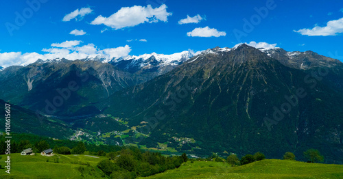 An expansive view of a majestic mountain range rising above a verdant green valley under an azure sky, casting dramatic shadows across the picturesque landscape.