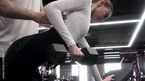 In the gym, a supportive male personal trainer provides guidance to a woman as she lifts a dumbbell. Athletic Support Male Personal Trainer Assisting Woman in Weightlifting