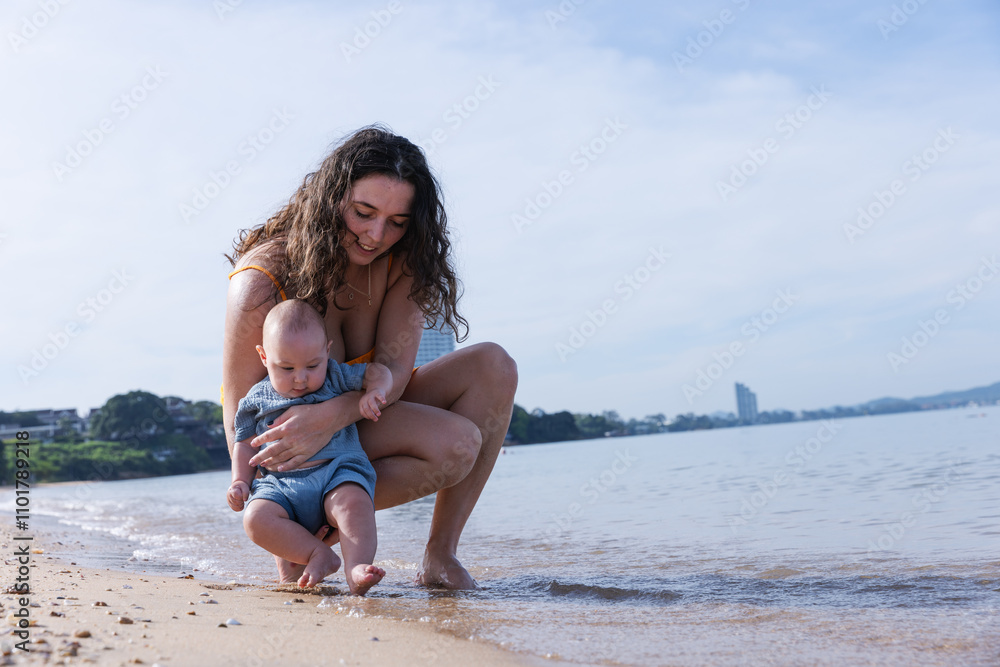 Naklejka premium Mother Gently Holding Baby by the Shoreline, Introducing Her Child to the Ocean Waves in a Relaxed Beach Environment, Sharing a Precious Moment in Nature on a Calm, Sunny Day