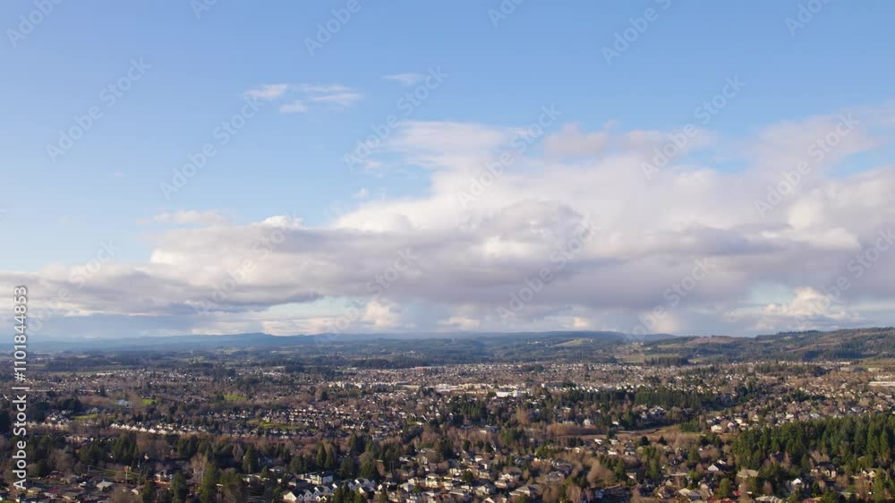 A breathtaking aerial view captures a sprawling suburban landscape under a vast blue sky, with soft, fluffy clouds drifting across the horizon and distant hills framing the scene.