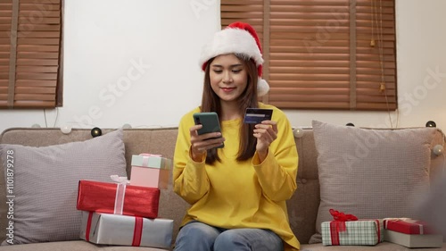 Woman wearing a santa hat is sitting on a couch surrounded by christmas presents and using her phone and credit card to shop online