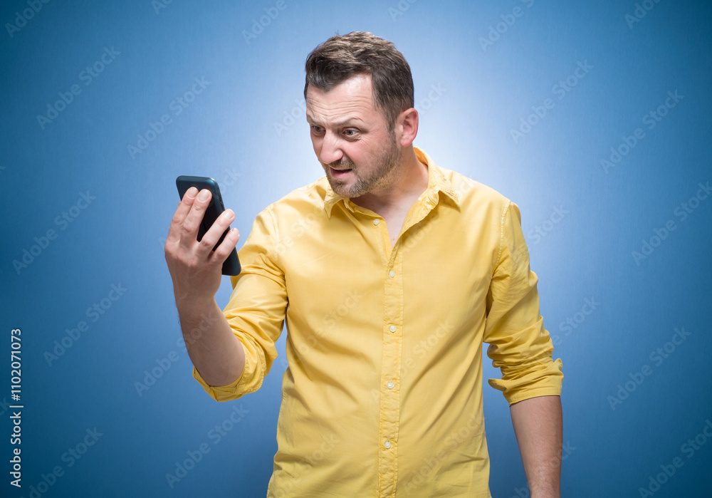 Upset young man looking to spoiled mobile phone over blue background, dresses in yellow shirt. Irritated guy with smartphone Upset young man looking to spoiled mobile phone over blue background, dresses in yellow shirt. Irritated guy with smartphone