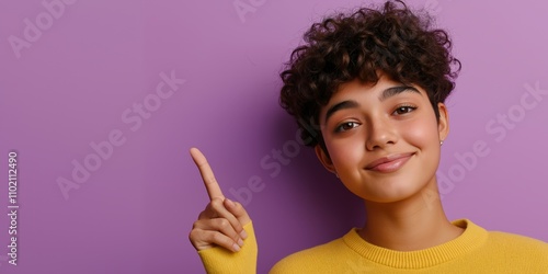 A modern and fresh studio image of a non-binary individual with short curly hair, wearing a casual sweater in pastel yellow, pointing upwards at copy space with an inviting smile