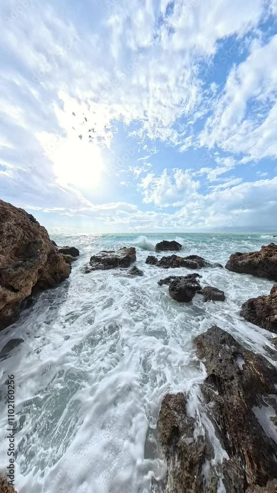 Vertical video. A wide-angle slow-motion view of foamy waves surging between jagged rocks, with the sun shining brightly and reflecting off the water under a picturesque sky. Vertical video. A wide-angle slow-motion view of foamy waves surging between jagged rocks, with the sun shining brightly and reflecting off the water under a picturesque sky.