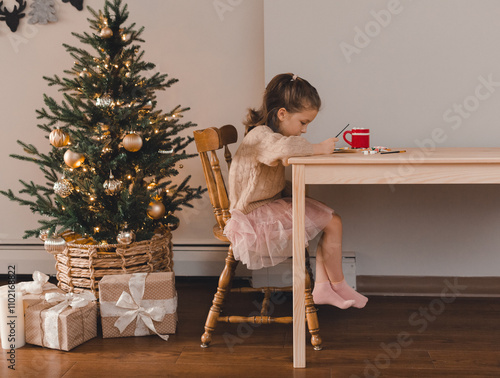 A little girl is sitting at a table drawing. A Christmas tree with gifts. Cozy Christmas atmosphere, home comfort