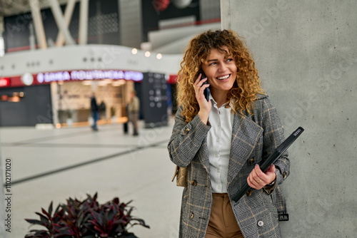 Amidst the railway station, a cheerful young woman in casual clothes walks, talks on the phone, and cradles a laptop, a delightful scene of multitasking and casual sophistication. photo