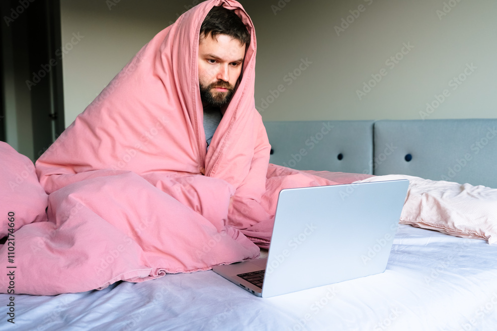 man wrapped in a pink blanket sits on a bed browsing a laptop, cozy at home, feel bad, sick or study in a comfortable home setting. man wrapped in a pink blanket sits on a bed browsing a laptop, cozy at home, feel bad, sick or study in a comfortable home setting.