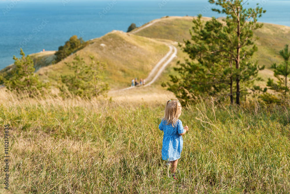 Cute little girl toddler in a dress walks on hills and paths. The concept of tourism with children, family travel. Hilly landscape with green meadows. Amazing view of rural scenery Cute little girl toddler in a dress walks on hills and paths. The concept of tourism with children, family travel. Hilly landscape with green meadows. Amazing view of rural scenery