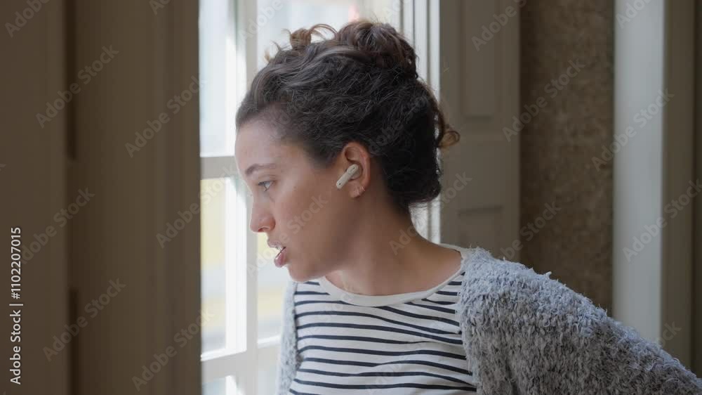 Woman standing next to window talking on phone using wireless headphones. Female businesswoman or student looking outside to street while having conversation over mobile phone using wireless earbuds Woman standing next to window talking on phone using wireless headphones. Female businesswoman or student looking outside to street while having conversation over mobile phone using wireless earbuds