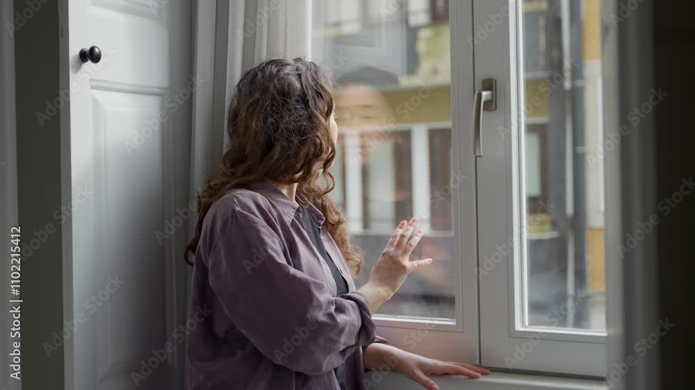 Sad unhappy woman stands by apartment window touching glass and looking outside. Female feeling lonely or depressed is waiting for someone or thinking and contemplating by city apartment window Sad unhappy woman stands by apartment window touching glass and looking outside. Female feeling lonely or depressed is waiting for someone or thinking and contemplating by city apartment window
