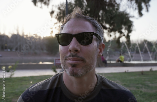 Portrait of a man in his 30s, wearing sunglasses sitting in the grass in the park at sunset. The white pergola columns in the background.	
