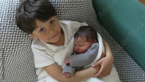 Young boy with a joyful expression holds his newborn sibling wrapped in a cozy blanket, showcasing a moment of pure happiness and brotherly love photo