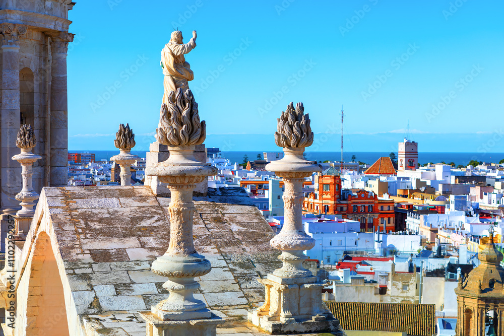 Jesus statue on top of Cathedral in Cadiz, Andalusia Spain. Sculpture is positioned on a rooftop, surrounded by ornate stone structures resembling flames. Panoramic city view with numerous buildings