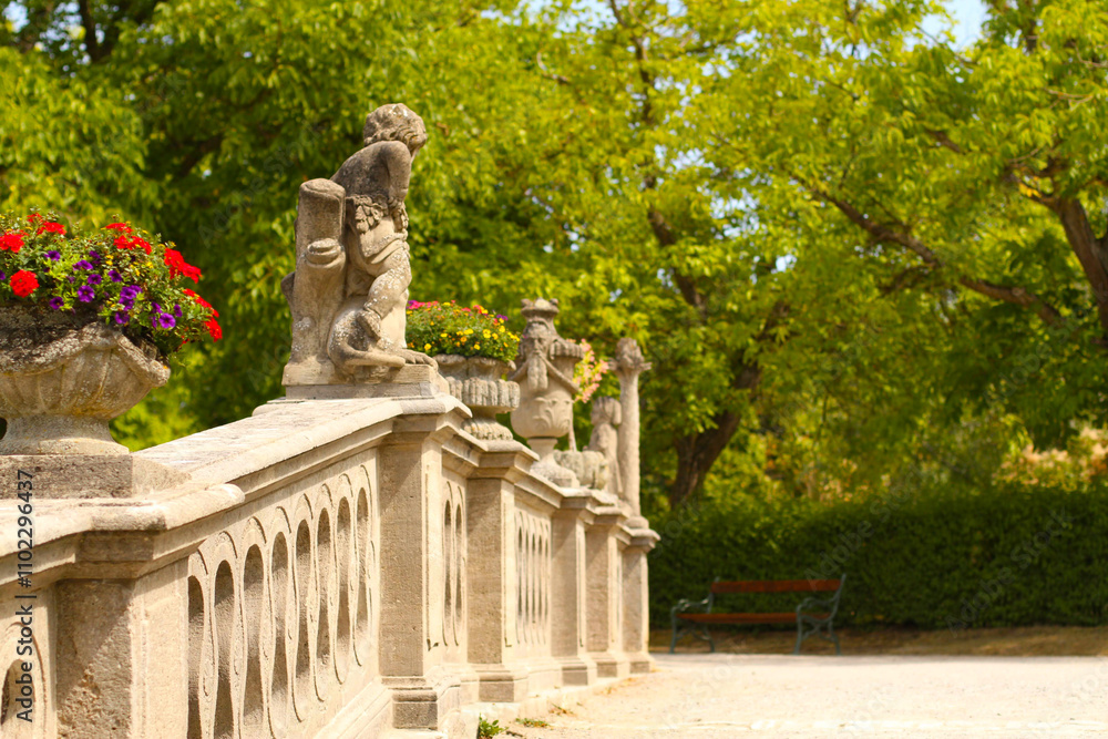 Obraz premium Eine elegante steinerne Balustrade mit kunstvollen Skulpturen und blühenden Blumenkübeln vor einer grünen Parklandschaft. Perfekte Harmonie von Architektur und Natur in einem sommerlichen Garten