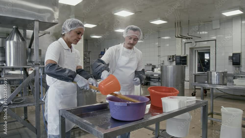 Medium full shot of two cheesemakers at work in factory – Caucasian woman holding wooden paddle, and male colleague pouring over hot water in tub with shredded curds for melting Medium full shot of two cheesemakers at work in factory – Caucasian woman holding wooden paddle, and male colleague pouring over hot water in tub with shredded curds for melting