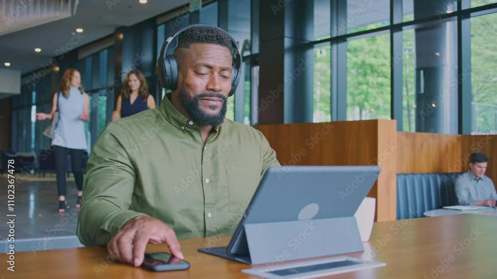 Man working on a tablet takes a break to look at his phone while sitting in a modern common area Man working on a tablet takes a break to look at his phone while sitting in a modern common area