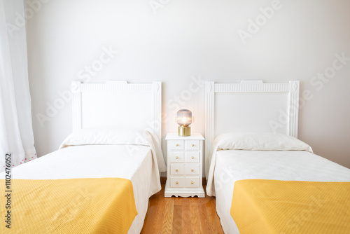 Bedroom with two small beds. Room with two 90cm beds made of white lacquered wood, white wall, natural wood floor, yellow bedspread, white lacquered drawer table, glass and navy gold lamp