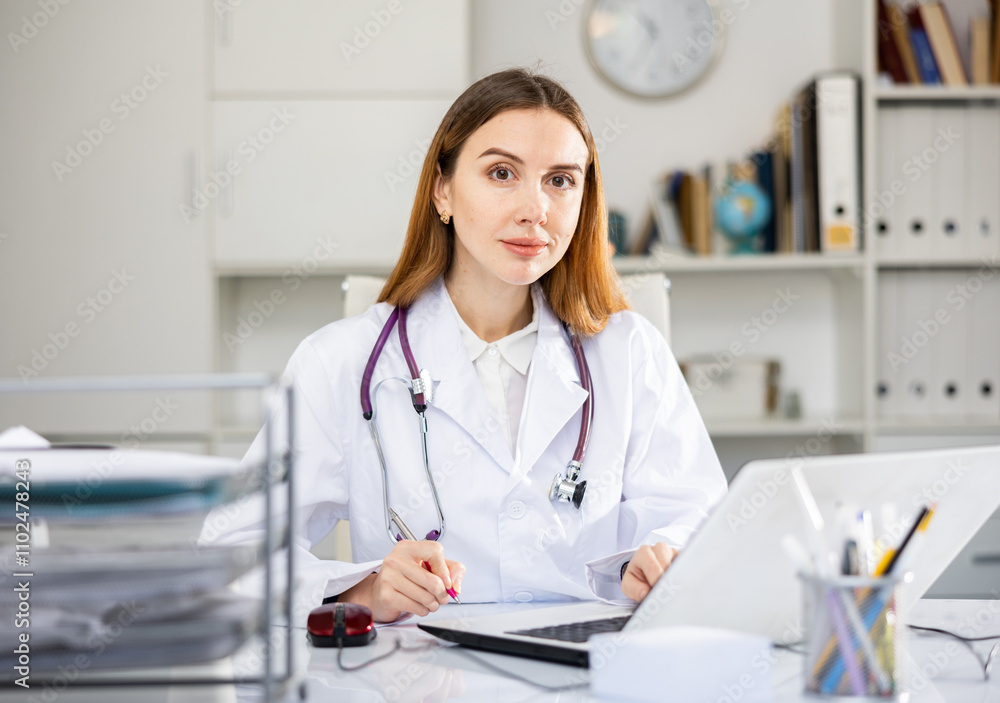 Young woman doctor sitting at table in her office and looking at camera. Young woman doctor sitting at table in her office and looking at camera.