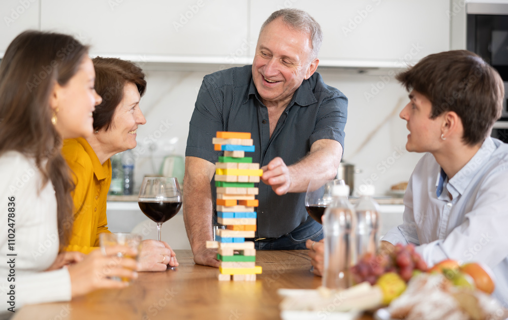 Elderly man engrossed in playing jenga with wife, adult son and daughter-in-law during fun family gathering with wine at home, concentrating on extracting block without collapsing tower Elderly man engrossed in playing jenga with wife, adult son and daughter-in-law during fun family gathering with wine at home, concentrating on extracting block without collapsing tower