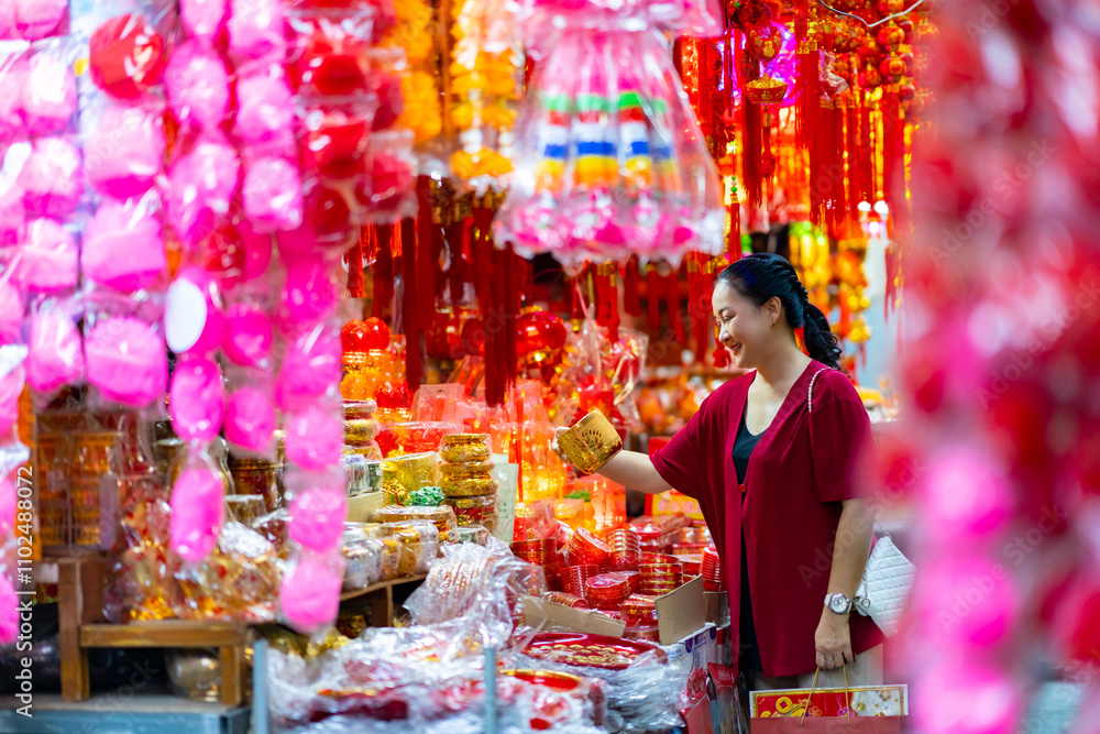 Obraz premium Happy Asian senior woman celebration Chinese lunar new year festival. Elderly retired woman in red blouse holding shopping bag walking and buying home decorative ornaments at Chinatown street market.