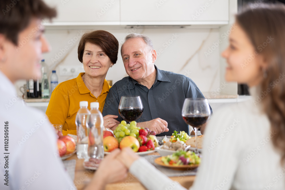 Couple sharing close moment at dining table, holding hands and locking eyes, while happy parents watching in background at family home. Blessing of parents Couple sharing close moment at dining table, holding hands and locking eyes, while happy parents watching in background at family home. Blessing of parents