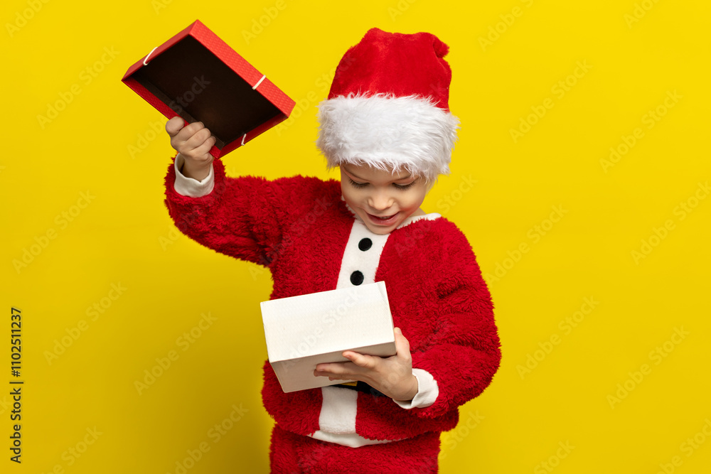 Excited child in a Santa costume opening a gift against a bright yellow background during the holiday season Excited child in a Santa costume opening a gift against a bright yellow background during the holiday season