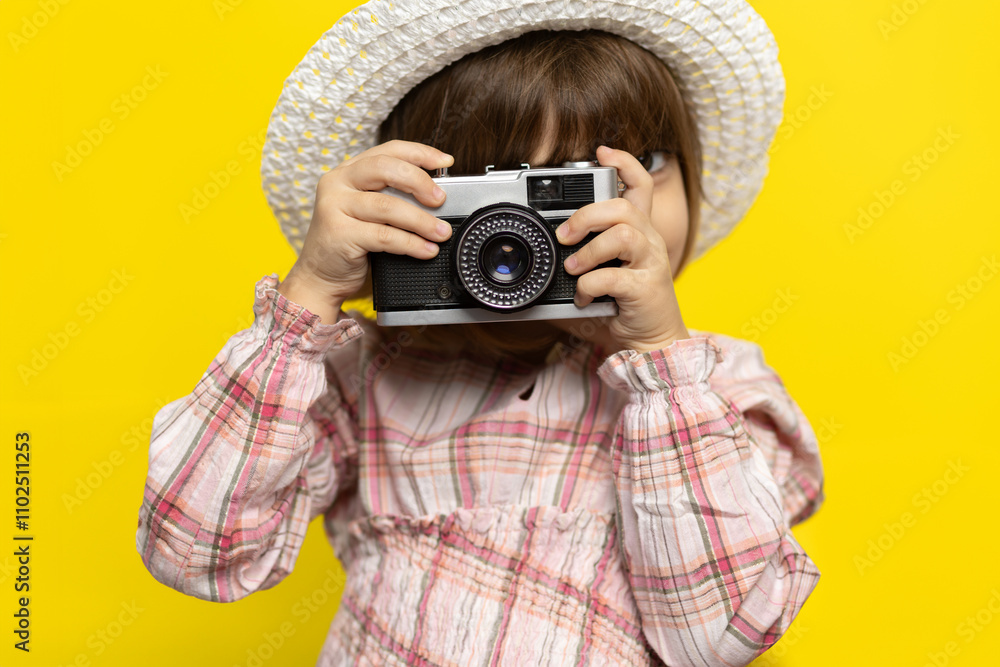 Young child in a straw hat holds a vintage camera against a vibrant yellow background while smiling joyfully Young child in a straw hat holds a vintage camera against a vibrant yellow background while smiling joyfully