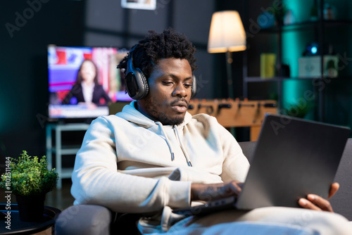 Cheerful man relaxing at home by watching TV series on laptop using headphones. Carefree person enjoying leisure time in RGB lit apartment at night, consuming entertainment media content