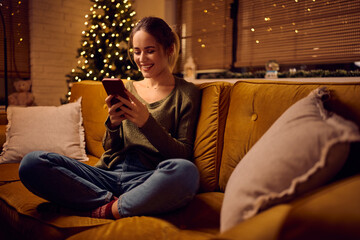 Happy woman texting on cell phone while relaxing on sofa on Christmas.