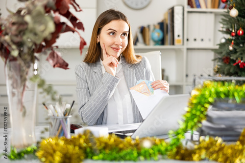 Portrait of positive young woman sitting at working place in office at New Year and receiving bonus money in envelope photo