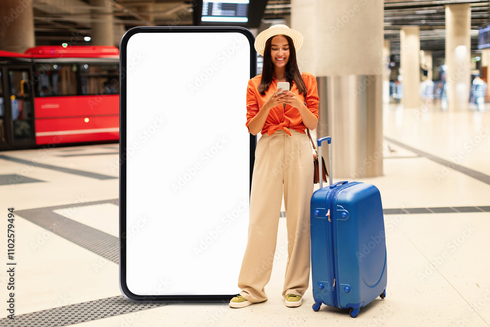 © Prostock-studio - In a bustling airport, a young woman stands beside a large blank advertisement display while checking her phone. She holds a suitcase, appearing ready to embark on her journey.