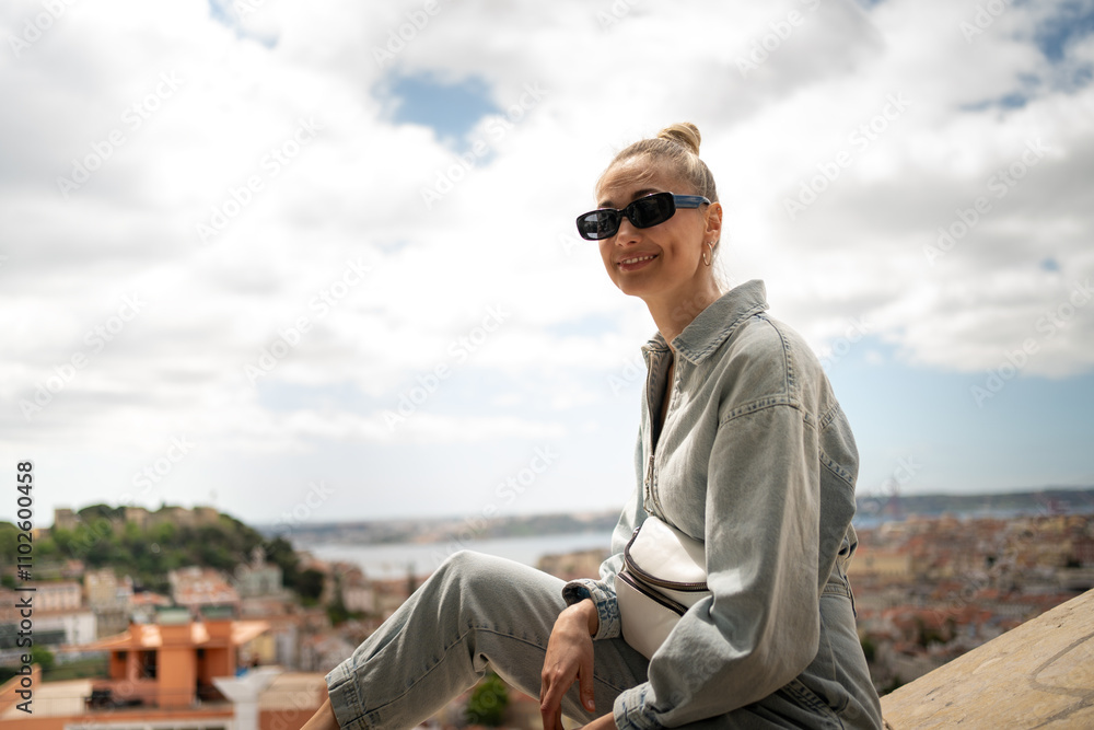 Happy female tourist sitting on retaining wall at Senhora do Monte in Lisbon viewpoint against cloudy sky. Portrait of smiling attractive woman in casualwear and sunglasses is on vacation. Happy female tourist sitting on retaining wall at Senhora do Monte in Lisbon viewpoint against cloudy sky. Portrait of smiling attractive woman in casualwear and sunglasses is on vacation.