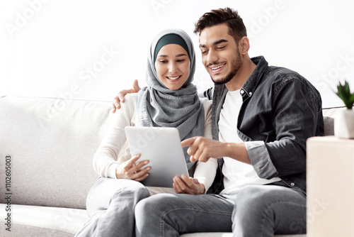 Beautiful middle-eastern couple sitting on couch with digital pad, happy arab man husband and wife woman in hijab moving to new house, buying furniture and electronics online via tablet, copy space photo