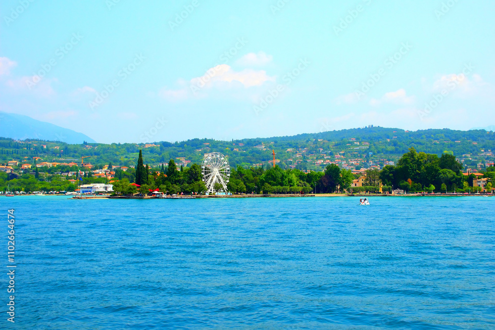 Interesting view at Bardolino on the eastern shore of Lake Garda with its white ferris wheel, the Lungolago Francesco Lenotti promenade, Beach Punta Cornicello, buildings, trees, vegetation, a boat Interesting view at Bardolino on the eastern shore of Lake Garda with its white ferris wheel, the Lungolago Francesco Lenotti promenade, Beach Punta Cornicello, buildings, trees, vegetation, a boat