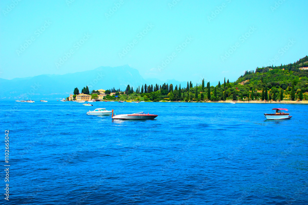 Ecstatic view at Punta San Vigilio with green cypresses and flourishing vegetation standing near human settlements, with different white motorboats skimming the calm rippling waters of Lake Garda Ecstatic view at Punta San Vigilio with green cypresses and flourishing vegetation standing near human settlements, with different white motorboats skimming the calm rippling waters of Lake Garda