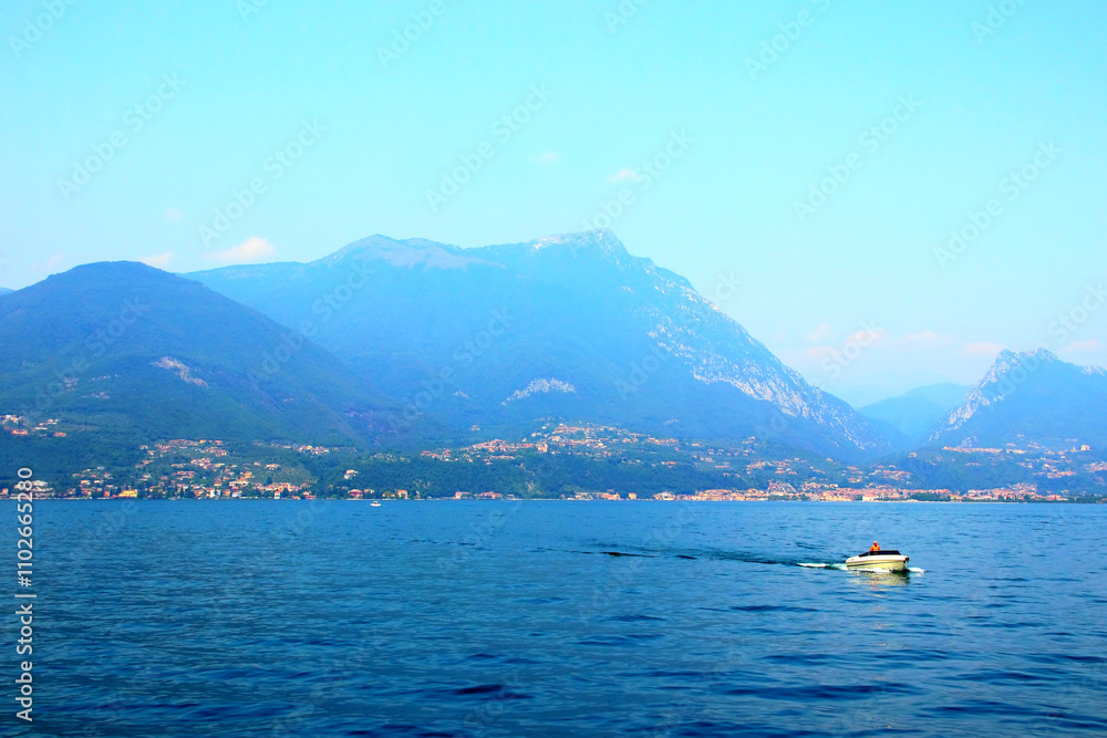 Impressive scenery on Lake Garda, majestic mountains in the hazy azure sky dominating over human settlements with many buildings perched on the underlying hills with vegetation, a cruising motorboat Impressive scenery on Lake Garda, majestic mountains in the hazy azure sky dominating over human settlements with many buildings perched on the underlying hills with vegetation, a cruising motorboat