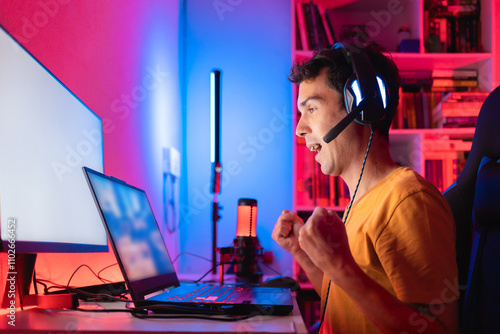 Gamer celebrating victory in an online multiplayer game match on a gaming laptop in his home room, illuminated by colorful LED lights, surrounded by streaming equipment and accessories photo