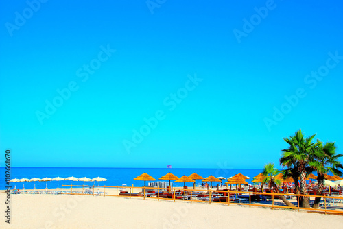 Paradisiac view at Civitanova Marche with gradient azure sky, swathes of blue Adriatic Sea, white and brown parasols, sandy beach, beach equipment, palm trees, an offshore structure visible in the sea