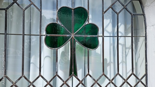 A stained-glass window featuring a green shamrock design, framed by black leaded lines, with light streaming through a white curtain in the background.