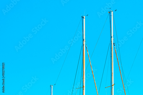 Upper parts of sailboat masts against a clear blue sky. Masts are equipped with various rigging lines and small devices at their tops, creating a visually scene due to the contrast with vibrant sky