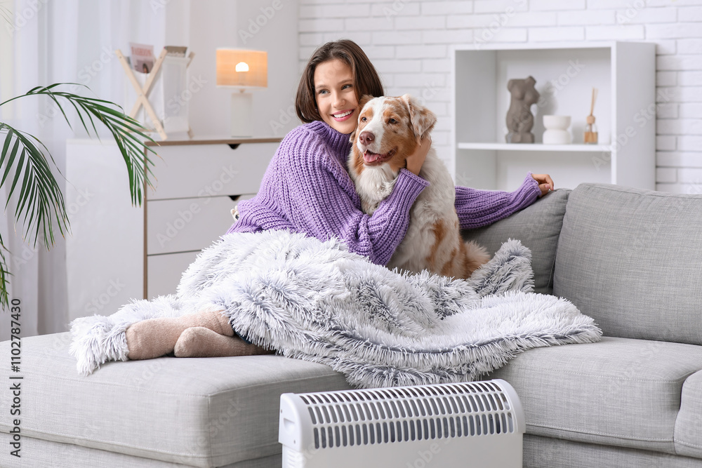 Young woman with blanket and Australian Shepherd dog sitting on sofa near heater at home Young woman with blanket and Australian Shepherd dog sitting on sofa near heater at home